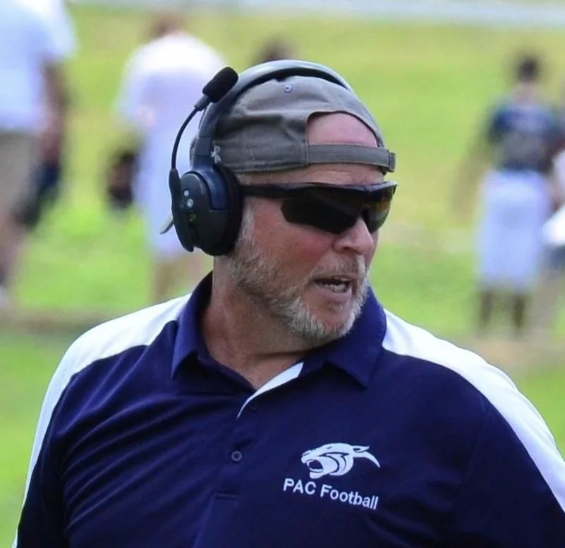 Sideline photograph of Coach Tim Youmans in a PAC Football polo, headset on and cap backwards — the reference photo used to engrave the plaque's portrait.